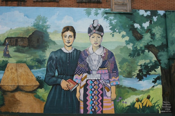 This snippet of the mural shows Laura Ingalls Wilder as a teacher next to a Hmong woman. To the left is the log bridge spanning Plum Creek, where the Ingalls family lived in a dug out.