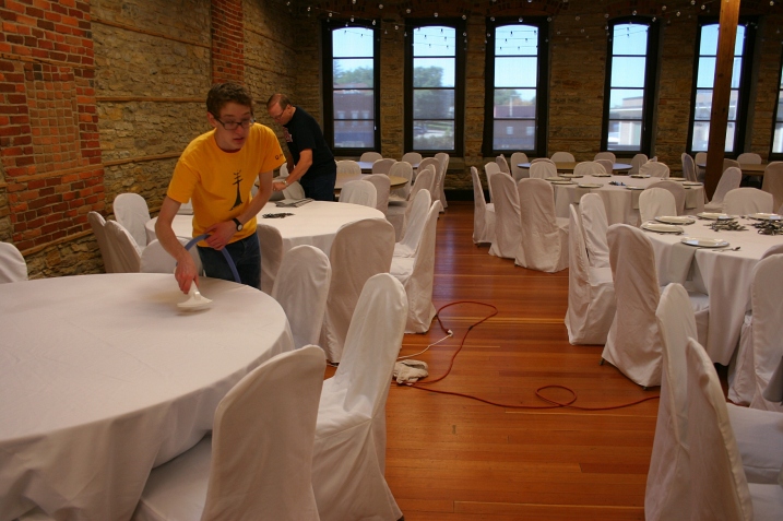 The bride's brother, Caleb, steam presses tablecloths while the father-of-the-groom, Eric, works on setting tables.