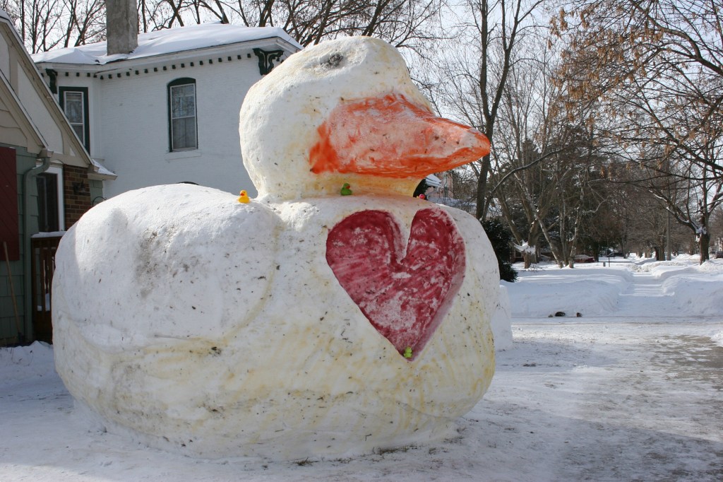 Multiple rubber duckies not sit atop the giant duck graced with a heart and a colored bill.