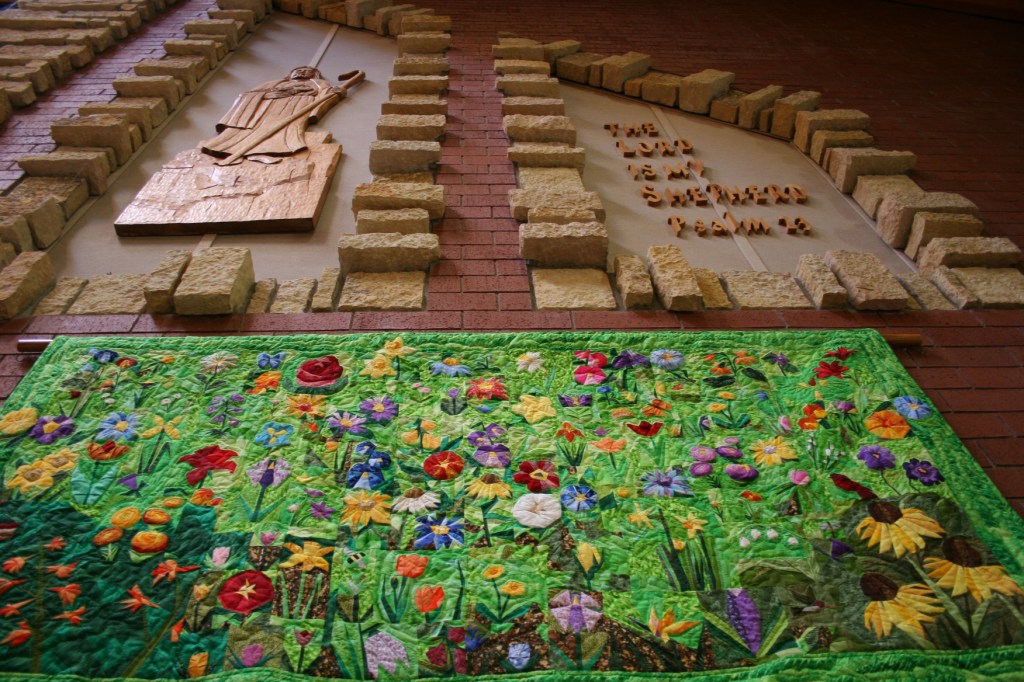 The beautiful and stunning floral quilt art by Kevin Kreger is displayed below a wood carving by noted Faribault artist Ivan Whillock. Kreger coordinated the "Consider the Lilies" floral display at Trinity.
