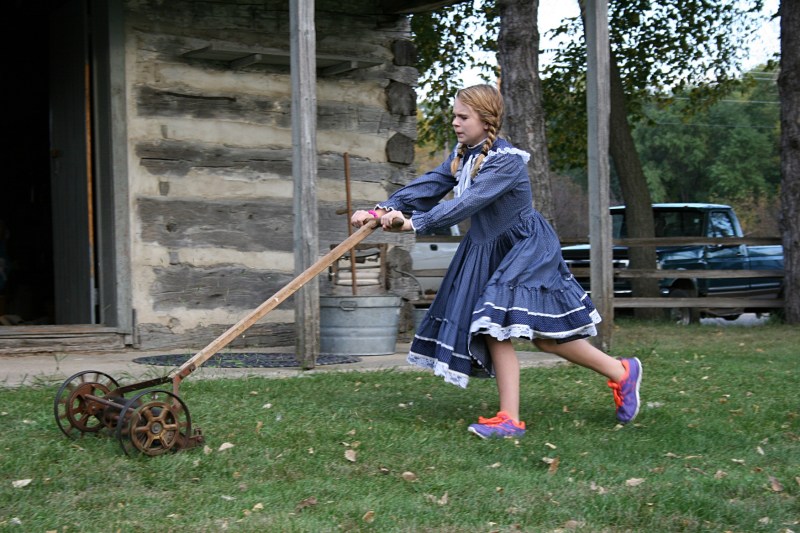 Kaylee, role-playing Katie, struggles to push an old-fashioned lawnmower across the lawn outside the log cabin.