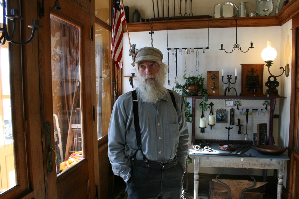 Blacksmith Tom Latane talks about his craft  in the front part of his shop. Behind him are examples of his work.