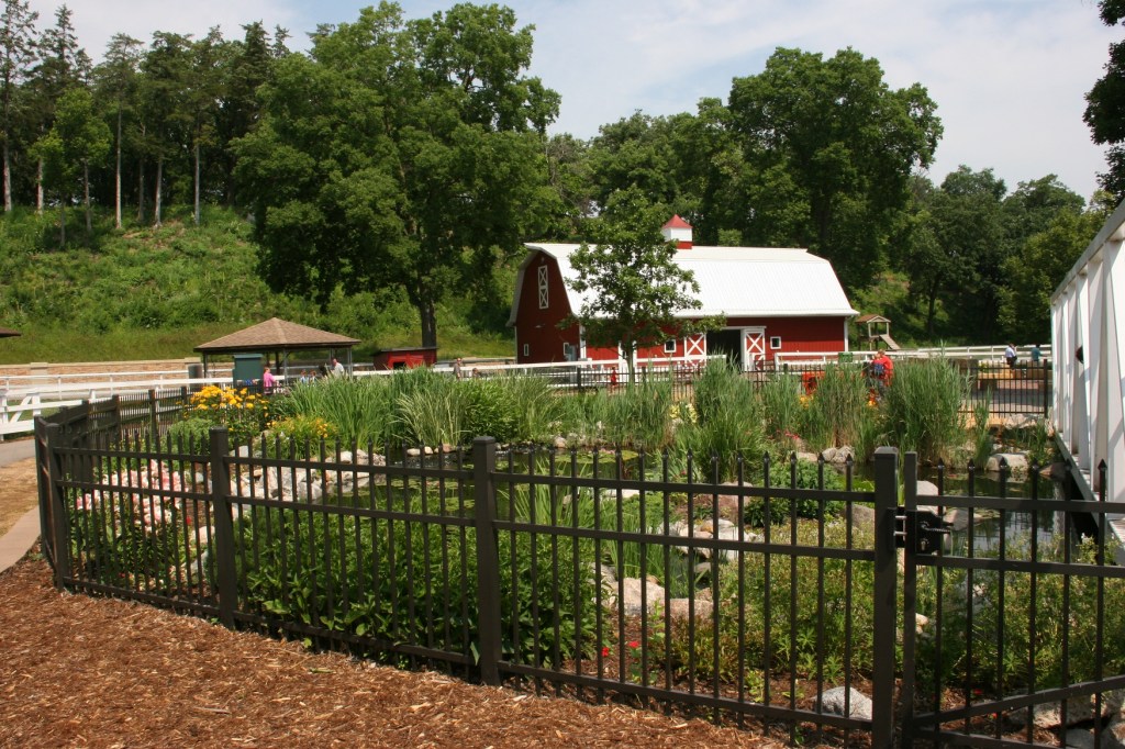 A fenced pond is in the foreground and a second barn type pole shed in the background.