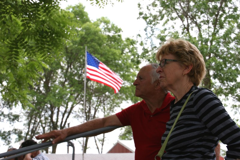 A couple listens to the music while sitting on portable bleachers under a canopy of trees.