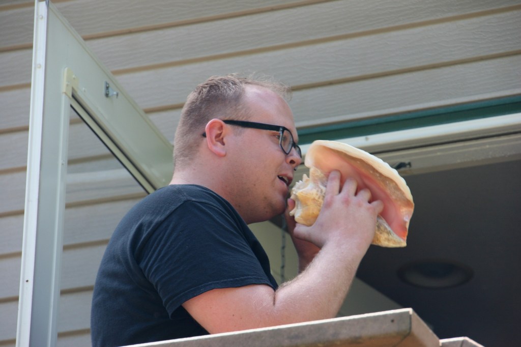 Justin stands atop a deck and calls the family to lunch by blowing into a conch shell.